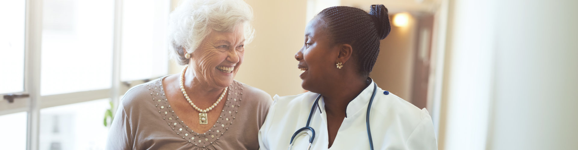 Senior woman walking in the nursing home supported by a caregiver. Nurse assisting senior woman.