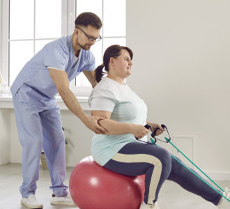 Nurse men helping fat patient to do sport stretching fitness exercises with rubber band sitting on fit ball in clinic. Male physiotherapist or health care worker helping overweight women in rehab.