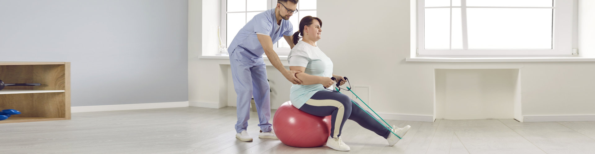 Nurse men helping fat patient to do sport stretching fitness exercises with rubber band sitting on fit ball in clinic. Male physiotherapist or health care worker helping overweight women in rehab.