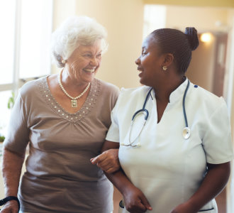 Senior woman walking in the nursing home supported by a caregiver. Nurse assisting senior woman.