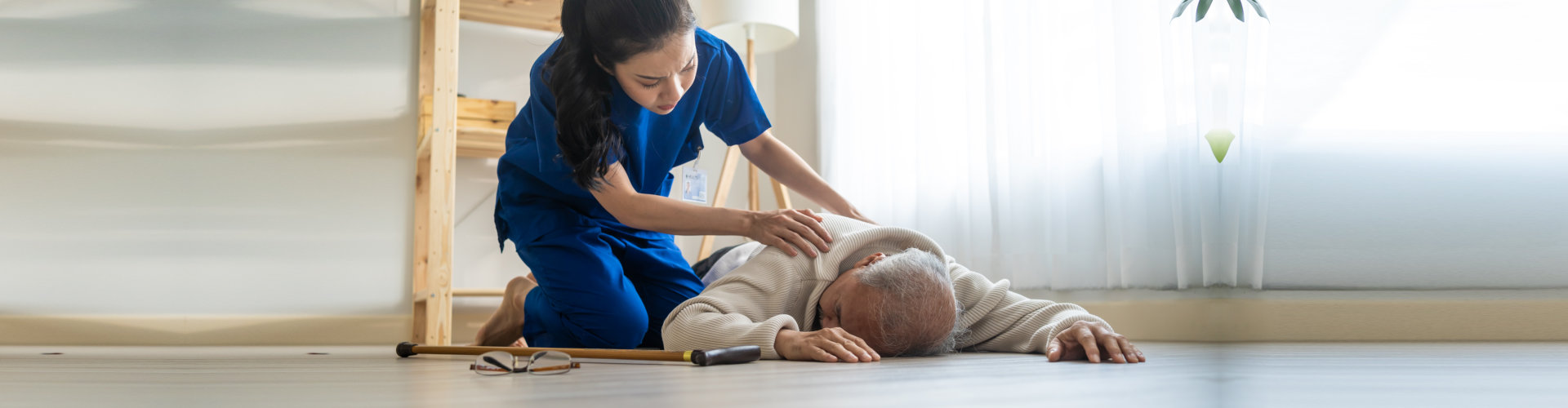 Asian caregiver helping senior male from falling on the ground at home. Elderly older patient having an accident after doing physical therapy then rescued by attractive therapist nurse in living room.